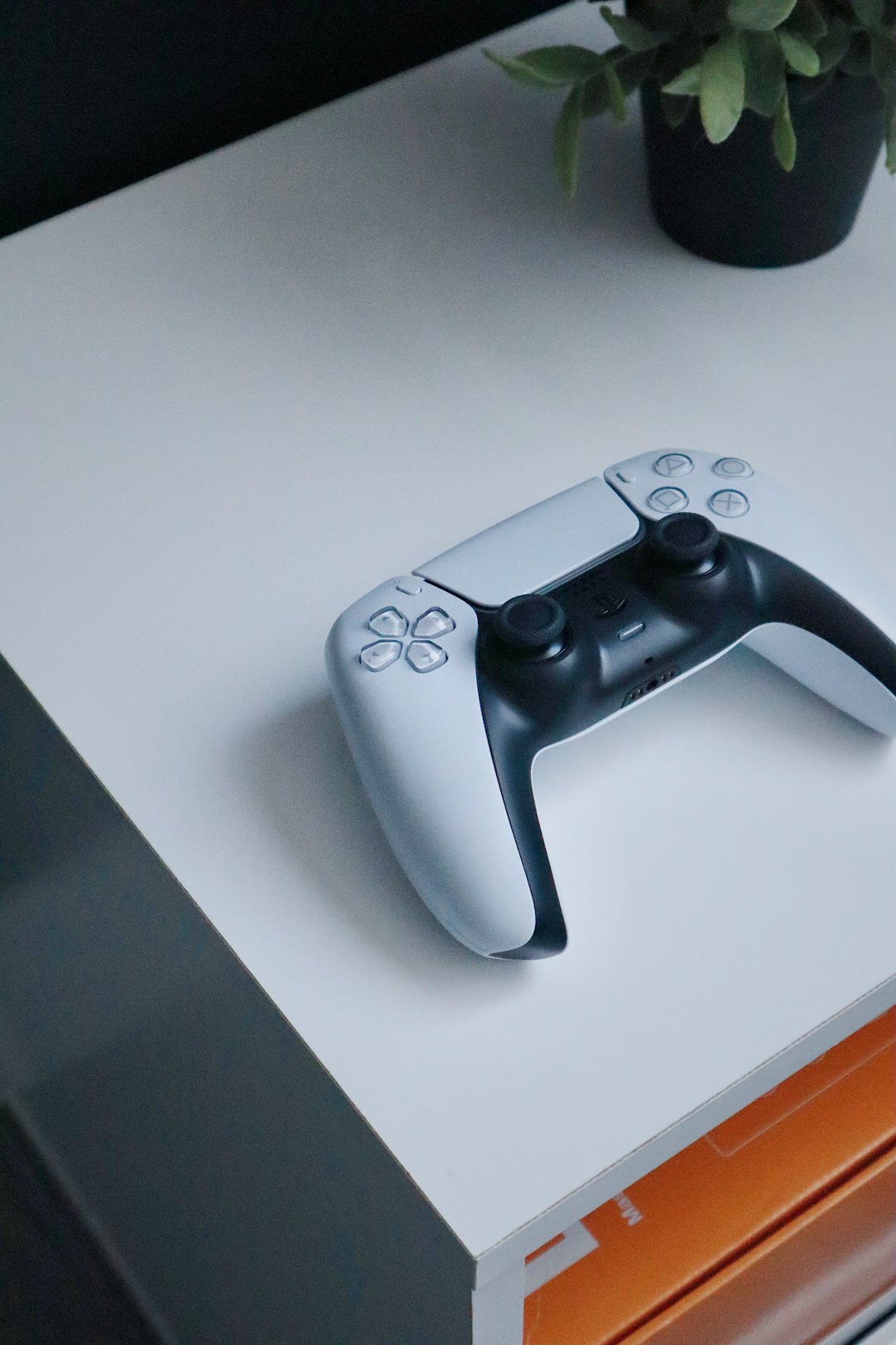 Playstation 5 (PS5) controller on a white night stand with an orange box in the bottom right and a plant in the top right. by Games Like white xbox one game controller