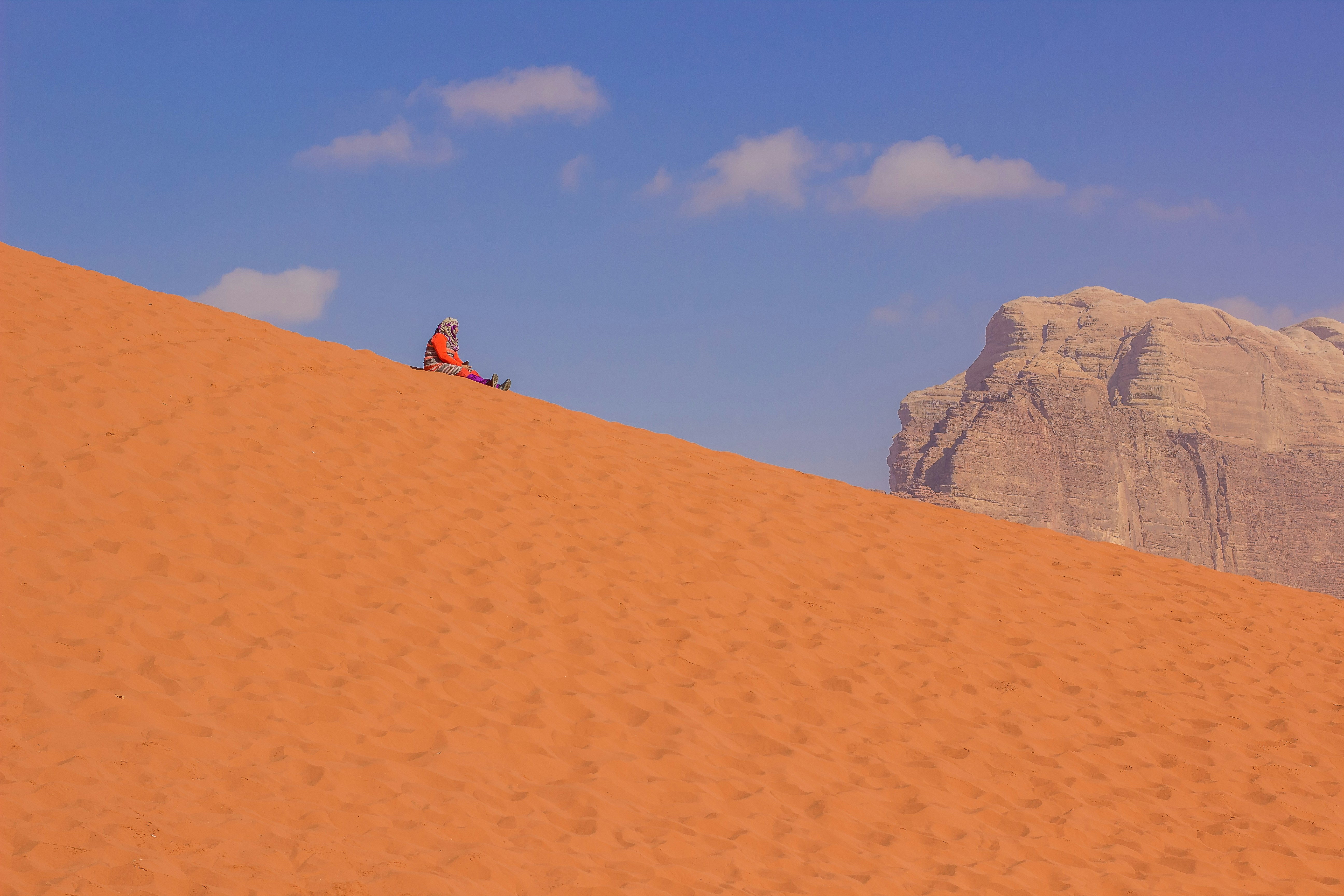 Funny travel photography of old woman sit on sand of desert dune hill and rest in Wadi Rum world famous travel site of Jordan Middle East region country, yellow background in clear weather day time by Games Like person in black jacket and blue denim jeans standing on brown sand during daytime